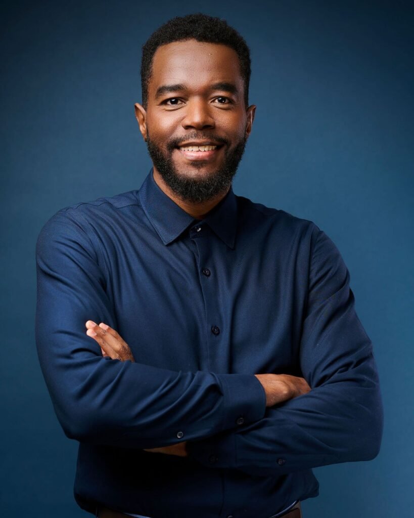 Berwin Sydney aujourd’hui, chemise marine, bras croisés, sourire, sur fond bleu