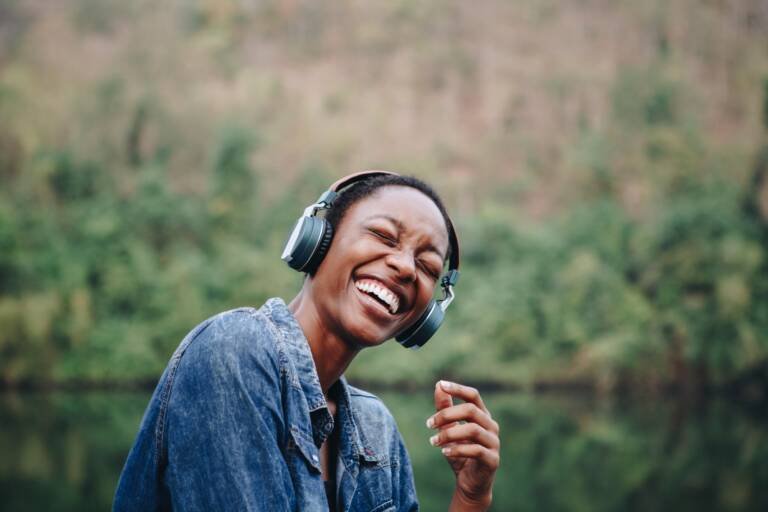 Femme noire souriante écoutant de la musique avec des écouteurs dans la nature, incarnant la puissance universelle de la musique.