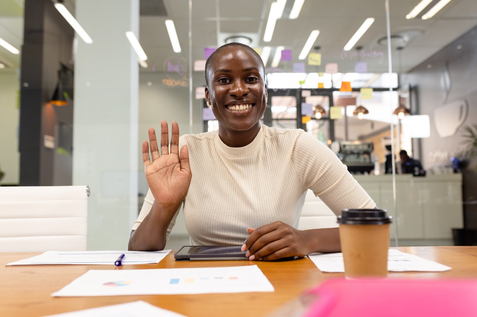 Femme noire souriante saluant la caméra dans un bureau moderne, assise devant des documents et une tablette, symbolisant une communicante d’OBNL