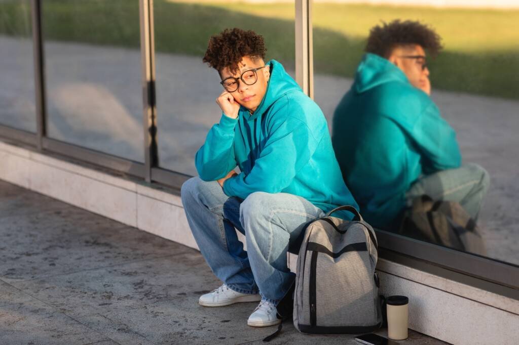 Young man sitting alone outside, reflecting, with his reflection in the glass — symbol of doubt, solitude, and inner questioning.
