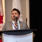 Conférencier en costume gris au podium, drapeaux canadien et francophone en arrière-plan lors d'un événement professionnel à Ottawa.