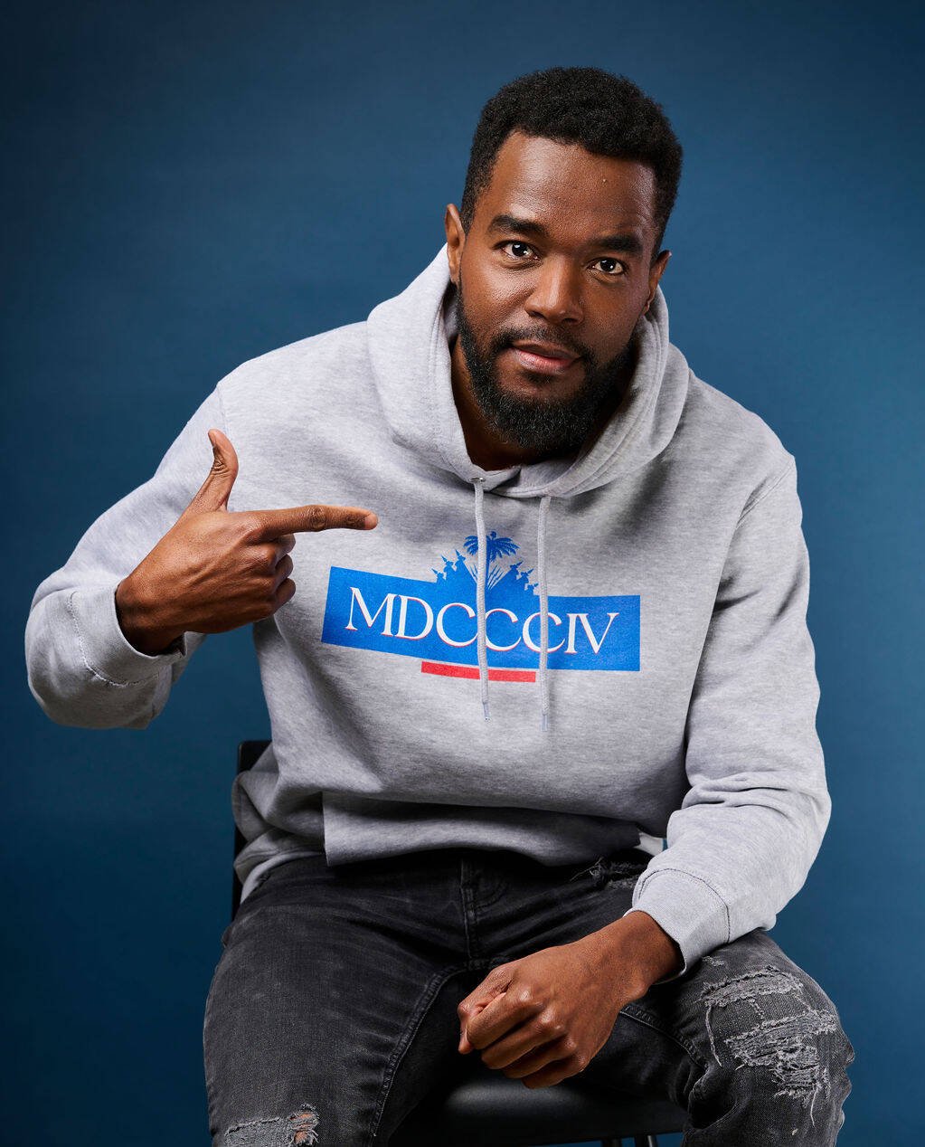Portrait de Berwin Sydney en hoodie gris pointant l’inscription MDCCCIV, en référence à 1804 et à l’indépendance d’Haïti, sur fond bleu.
