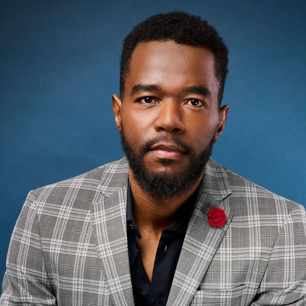 Portrait professionnel de Berwin Sydney en costume gris à carreaux, assis devant un fond bleu, les mains croisées.