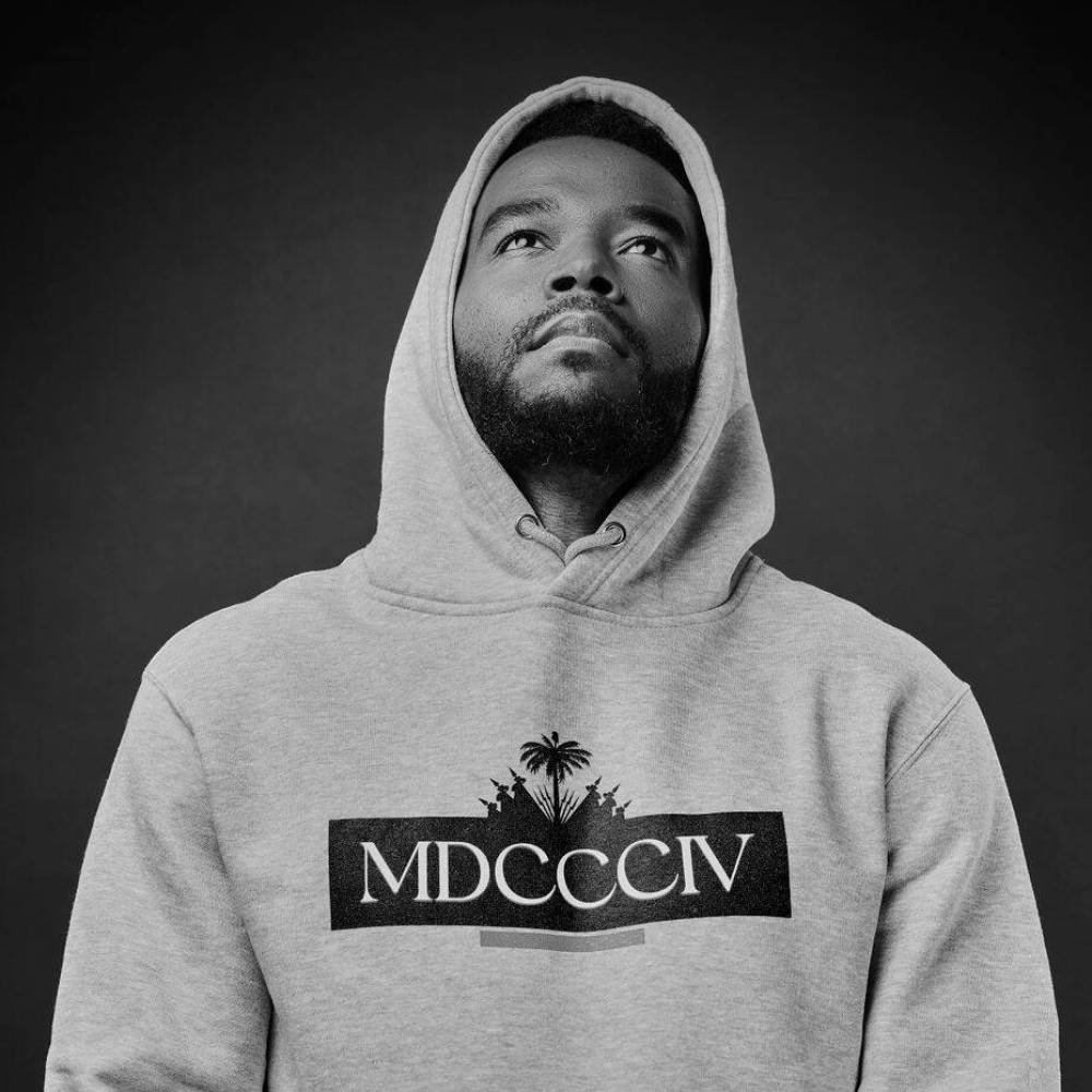 Portrait noir et blanc de Berwin Sydney en hoodie gris, regard levé, sur fond sombre.